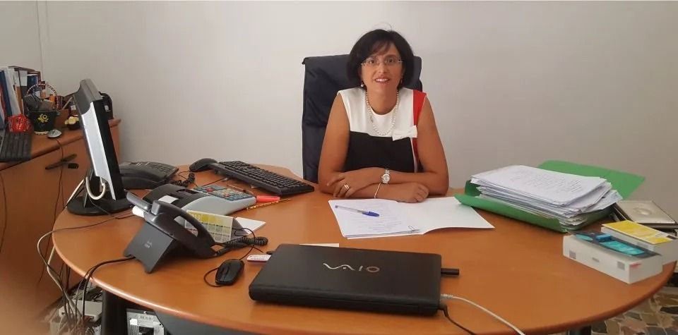Una donna seduta a una scrivania con documenti, computer portatile, telefono e pile di libri in un ufficio.