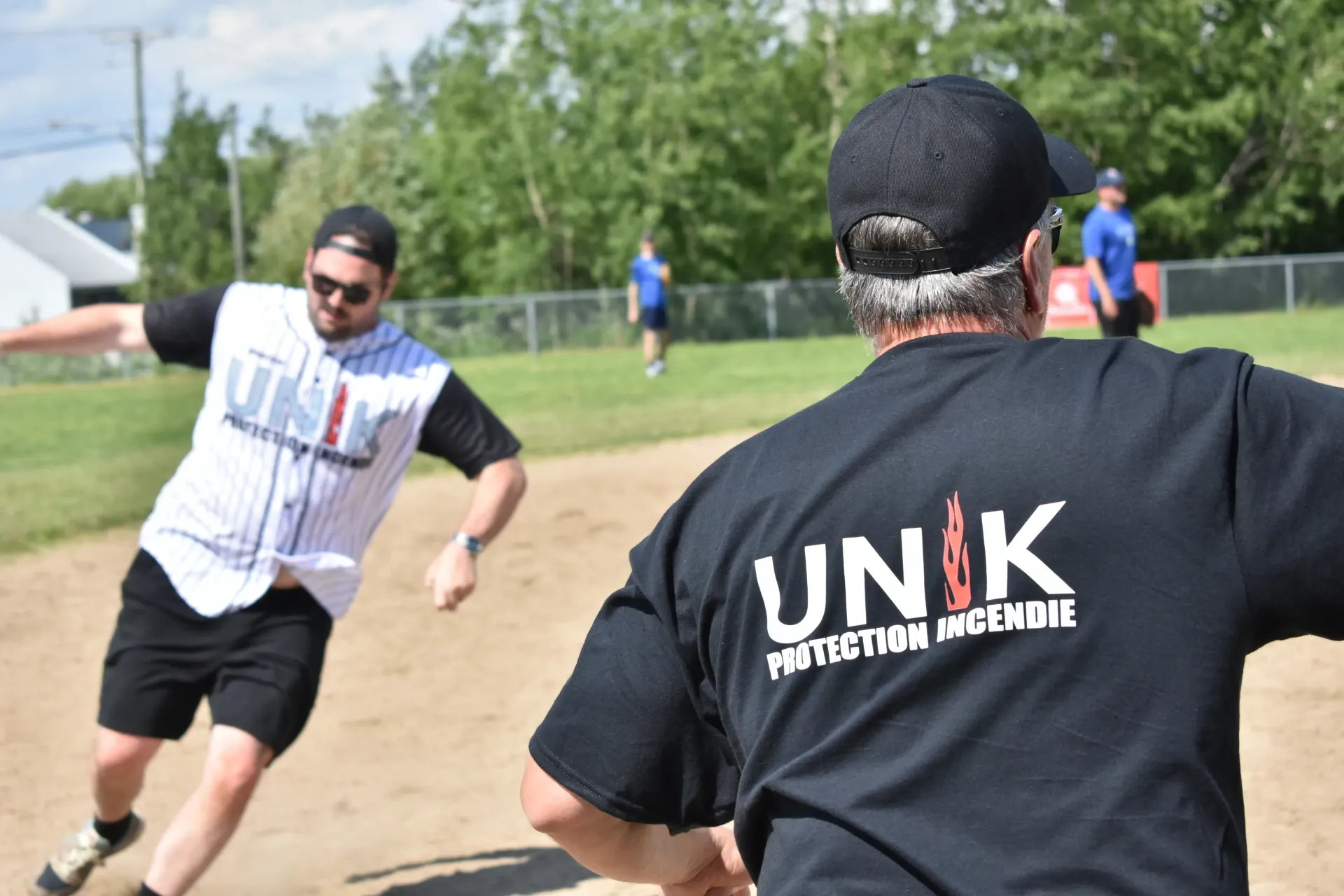 Deux personnes jouent au softball sur un terrain ensoleillé vêtue d'un t-shirt à l'effigie de UNK.