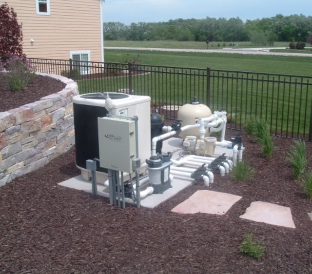 Pool equipment pad with a heater, pump, filter, and plumbing, surrounded by mulch and a fence.