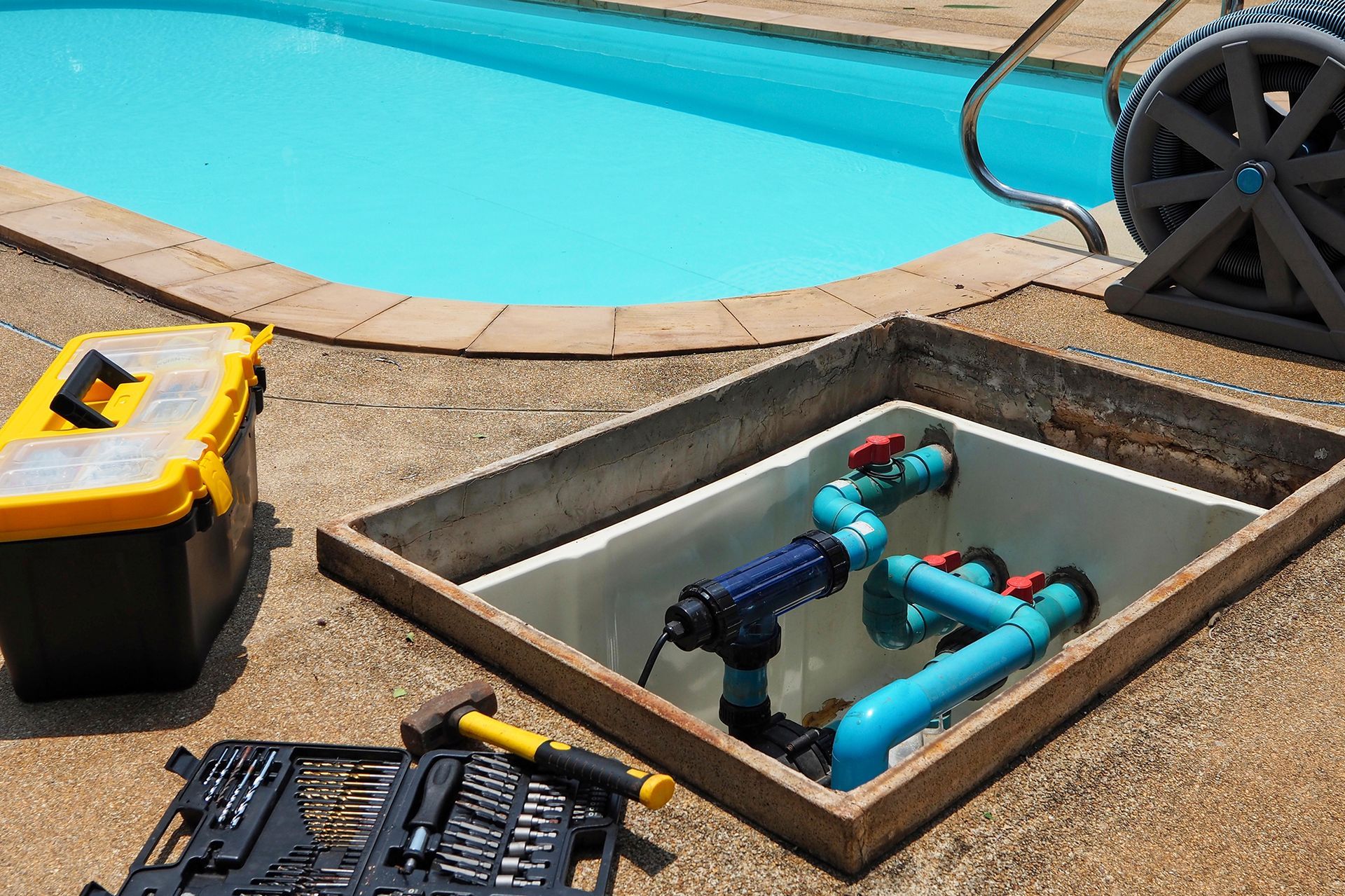 Pool equipment repair: open concrete pit with blue pipes, toolbox, and tools on a pool deck.