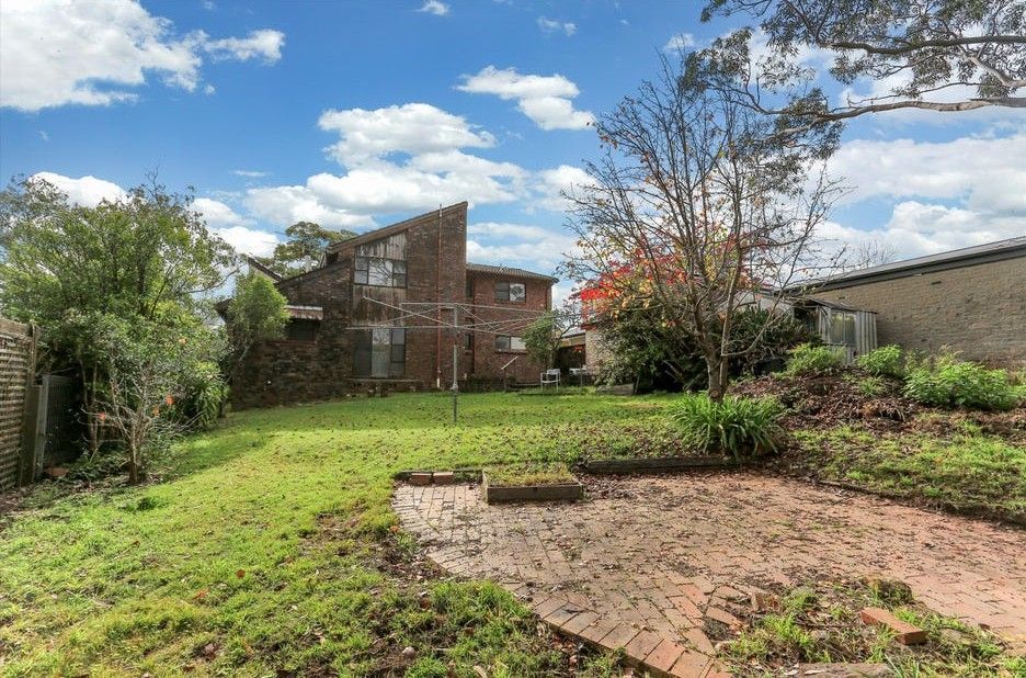 Backyard garden with brick patio, grassy lawn, shrubs, and a brick house under a partly cloudy sky
