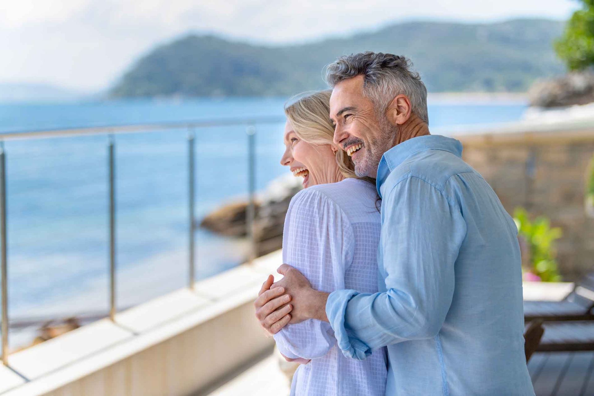A man and a woman are hugging each other on a balcony overlooking the ocean.
