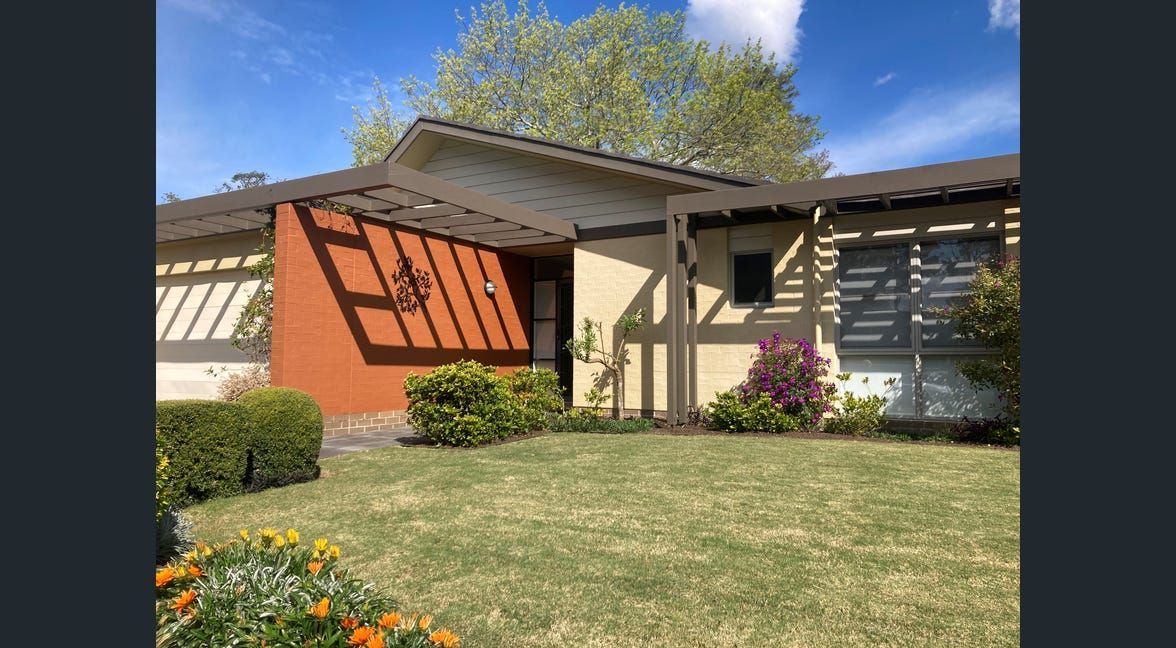 Sunlit house entrance with orange shutters, white columns, and a green lawn with flowers