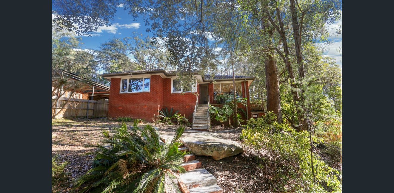 Small red cabin with a porch in a wooded yard, sunlight filtering through trees.