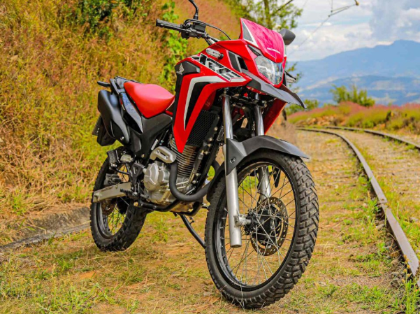 Red and black dual-sport motorcycle parked beside railway tracks on a dirt path in a hilly landscape