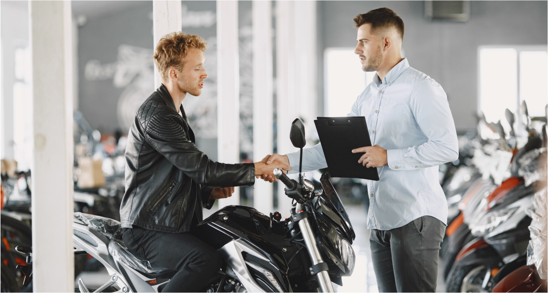 Um homem está sentado em uma motocicleta apertando a mão de um homem em um showroom de motocicletas.