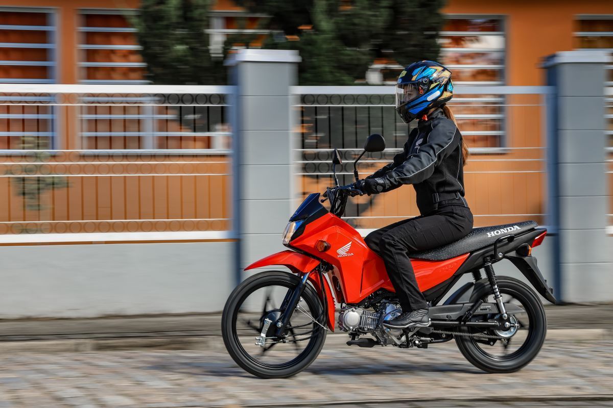 Motorcyclist in a blue helmet rides a red bike past a fence on a city street