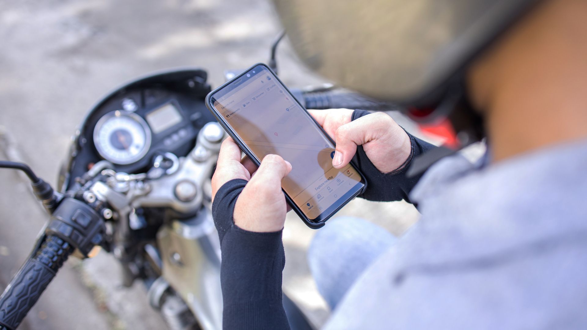 Person using a smartphone while seated on a motorcycle, with dashboard visible and a road in the background