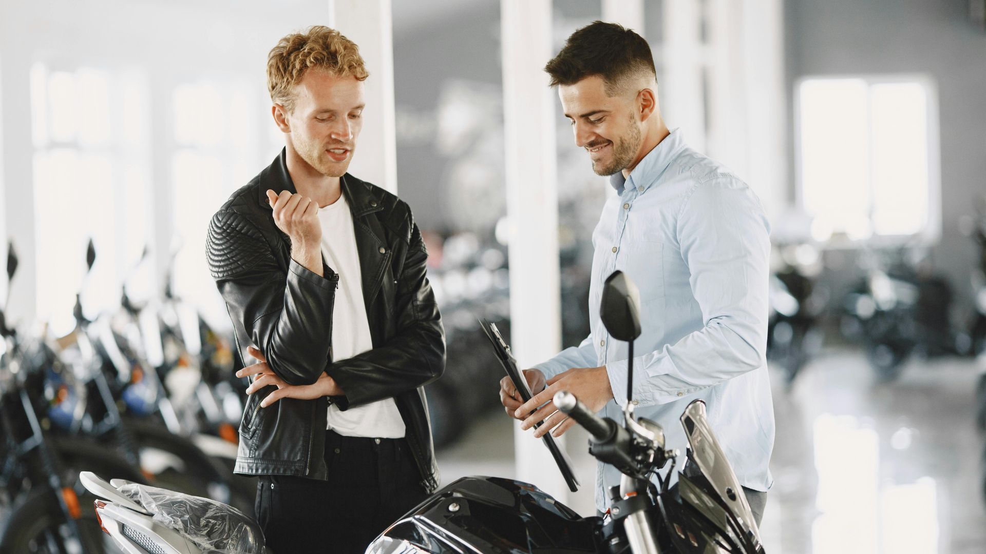 Two men discussing a motorcycle in a bright showroom, one gesturing while the other smiles.
