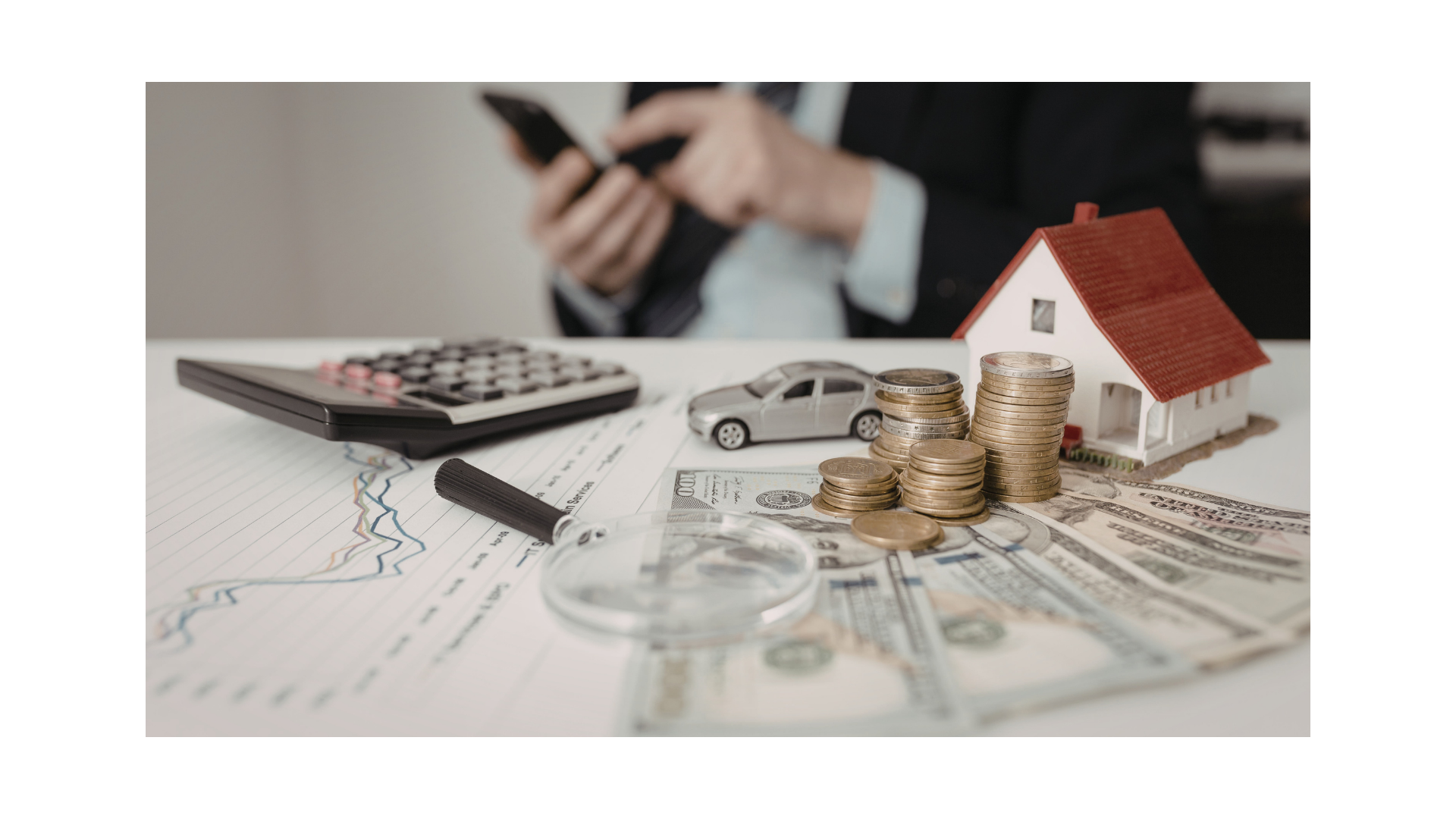 Calculator, coins, model house, and car on financial documents with a blurred person in the background