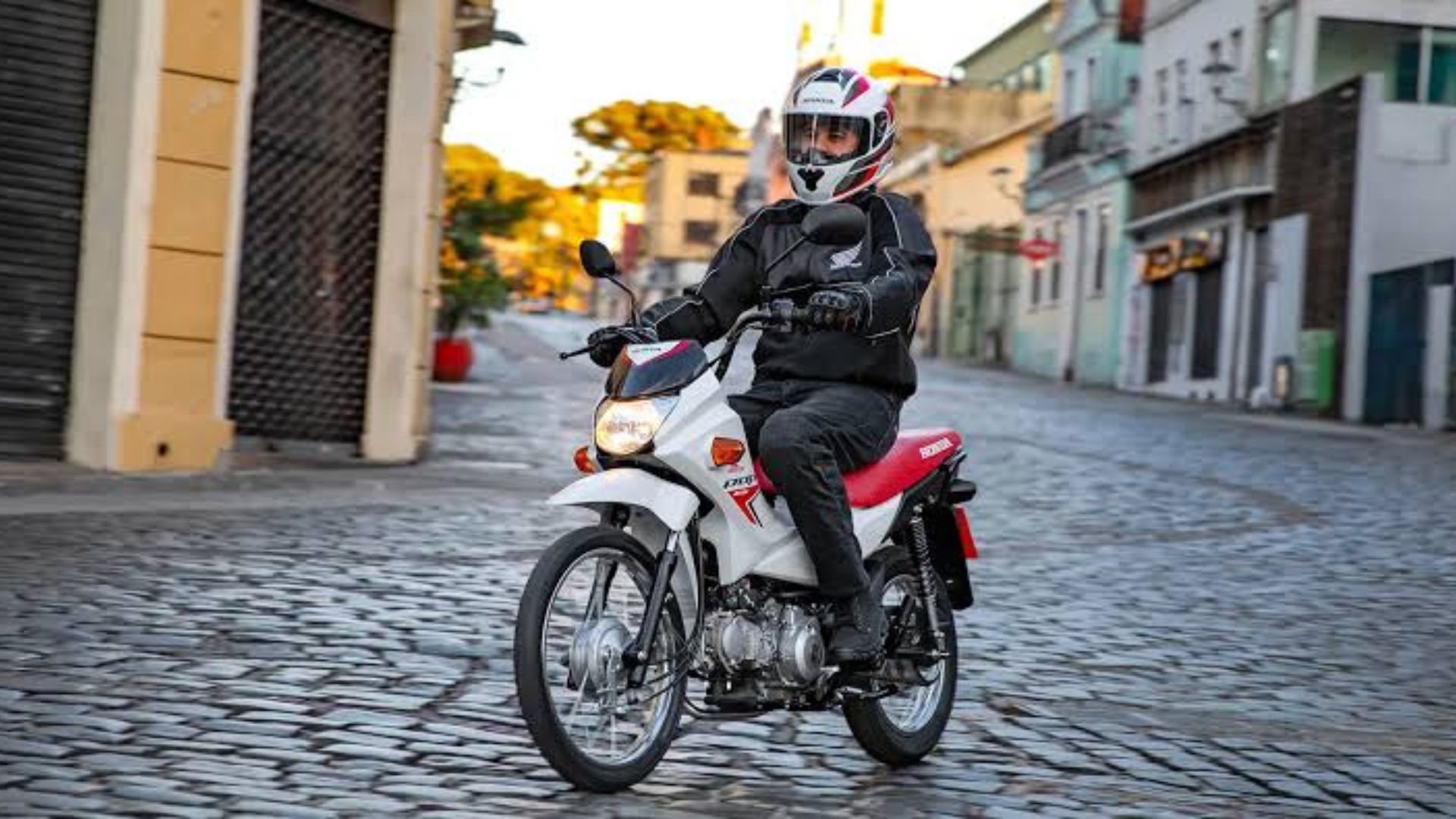 Motorcyclist in black gear riding a white scooter on a cobblestone street at sunset