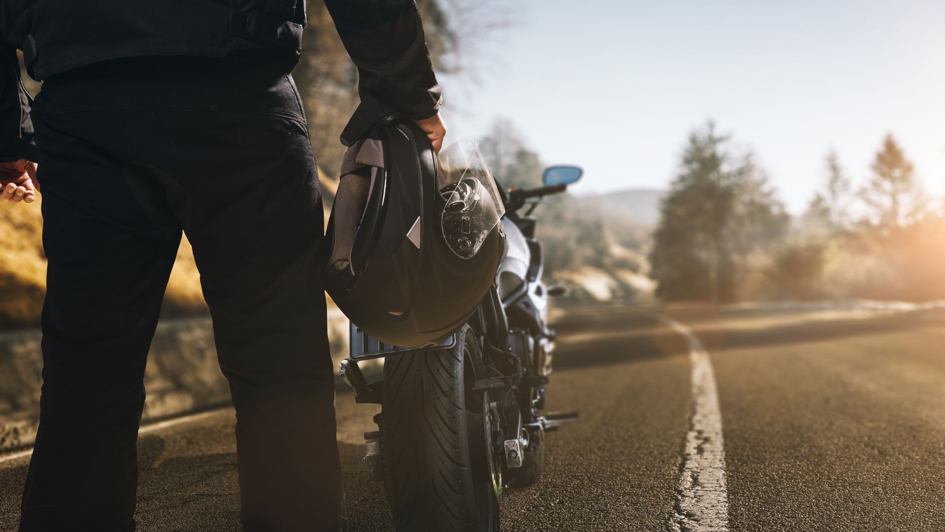 Person carrying a helmet beside a motorcycle on a sunlit road at dusk