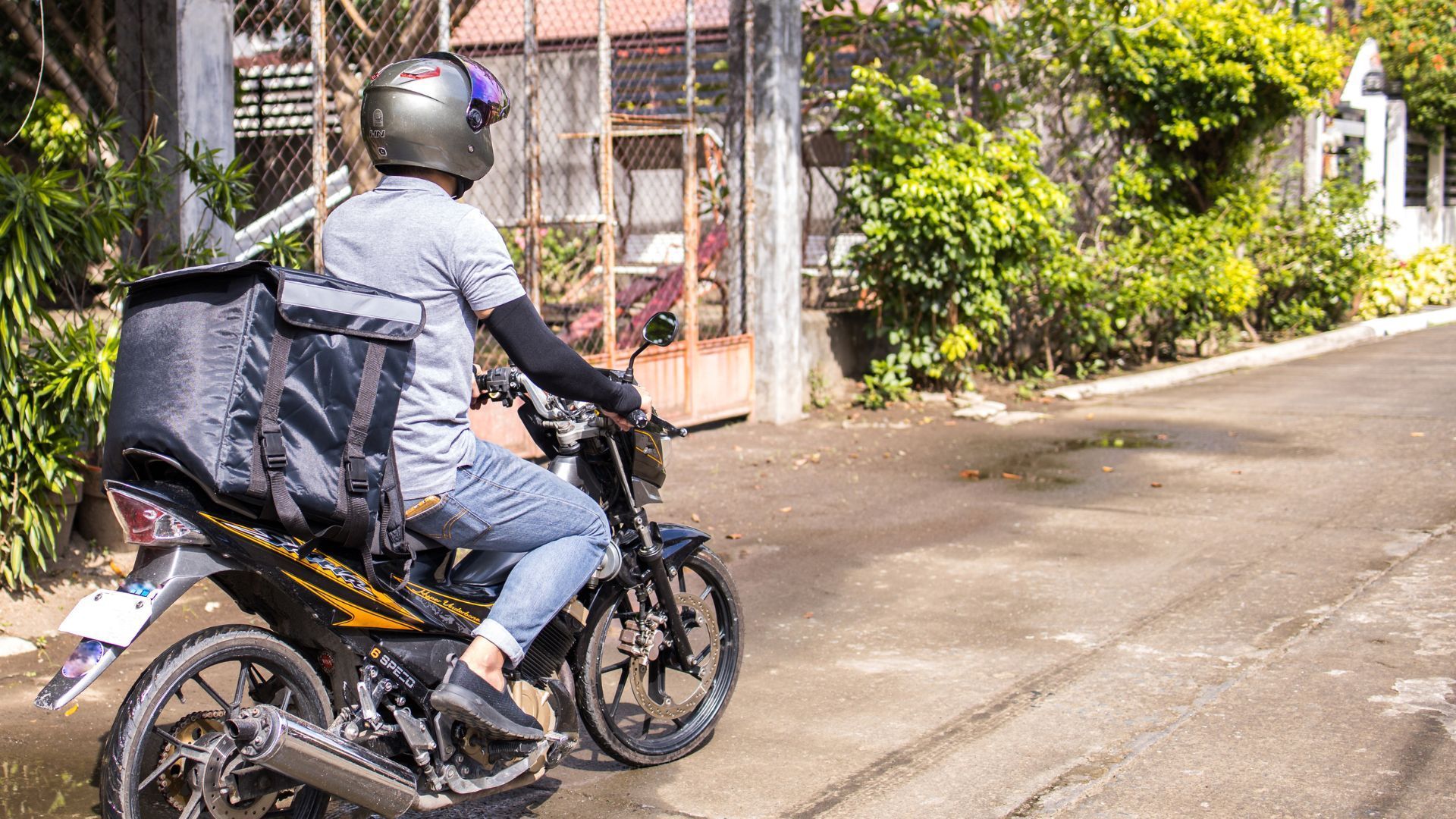 Motorcyclist in gray gear riding a black bike on a sunny residential street, carrying a large black cargo bag.