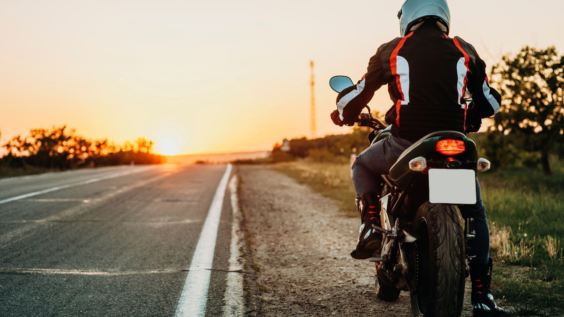 Motorcyclist paused on roadside at sunset, facing an empty highway