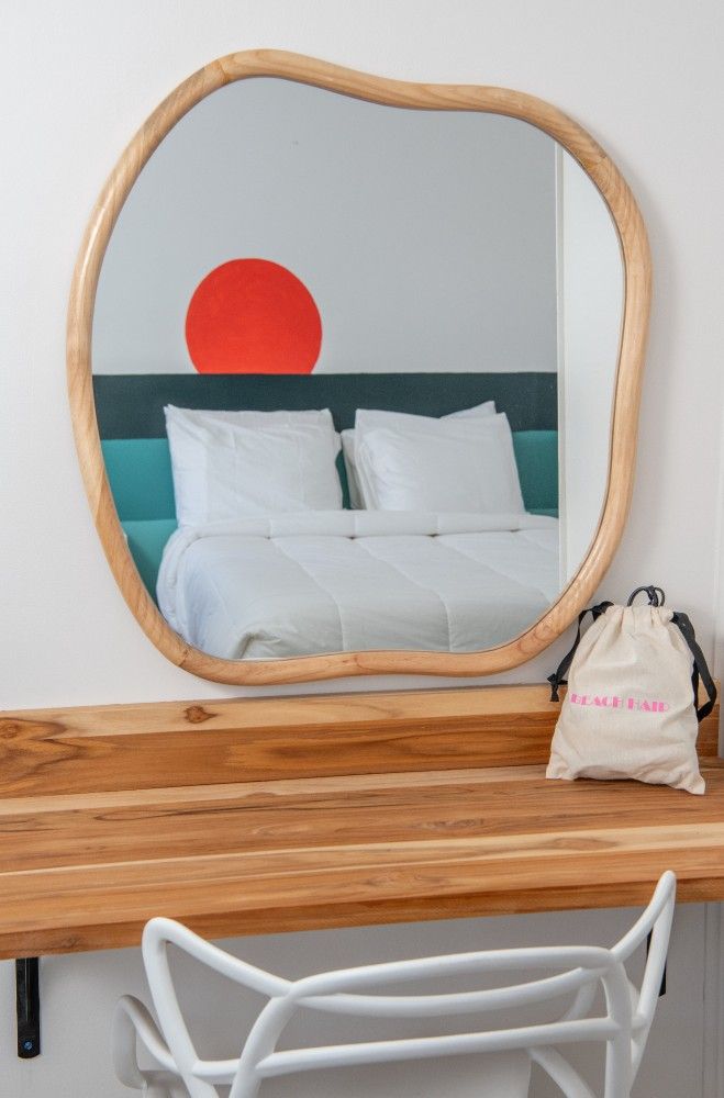 Wooden desk with wavy mirror reflecting a bed and red wall art, with a white chair and tote bag nearby