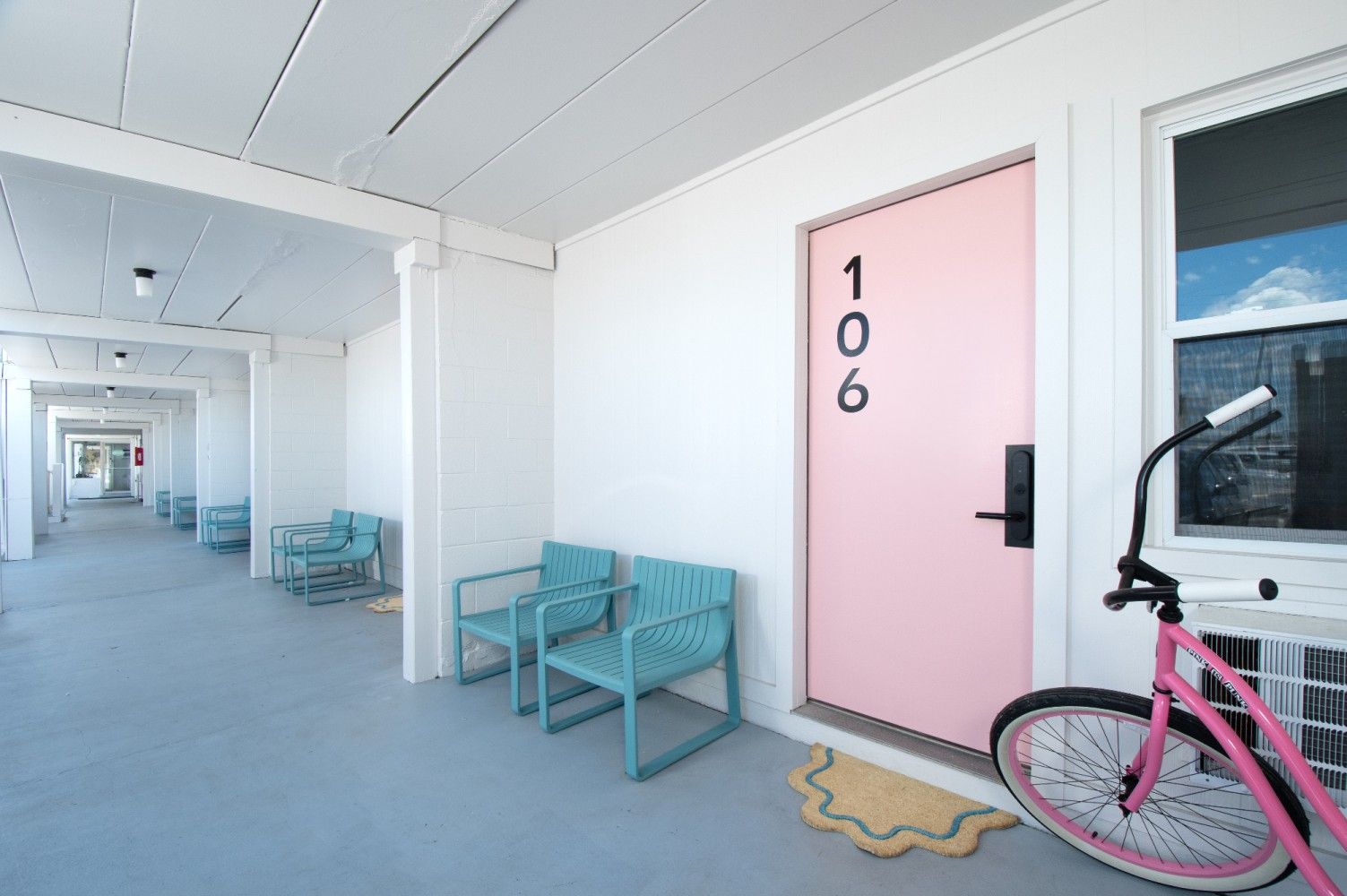 Pink door marked 106 beside turquoise chairs in a bright white hallway, with a pink bicycle nearby