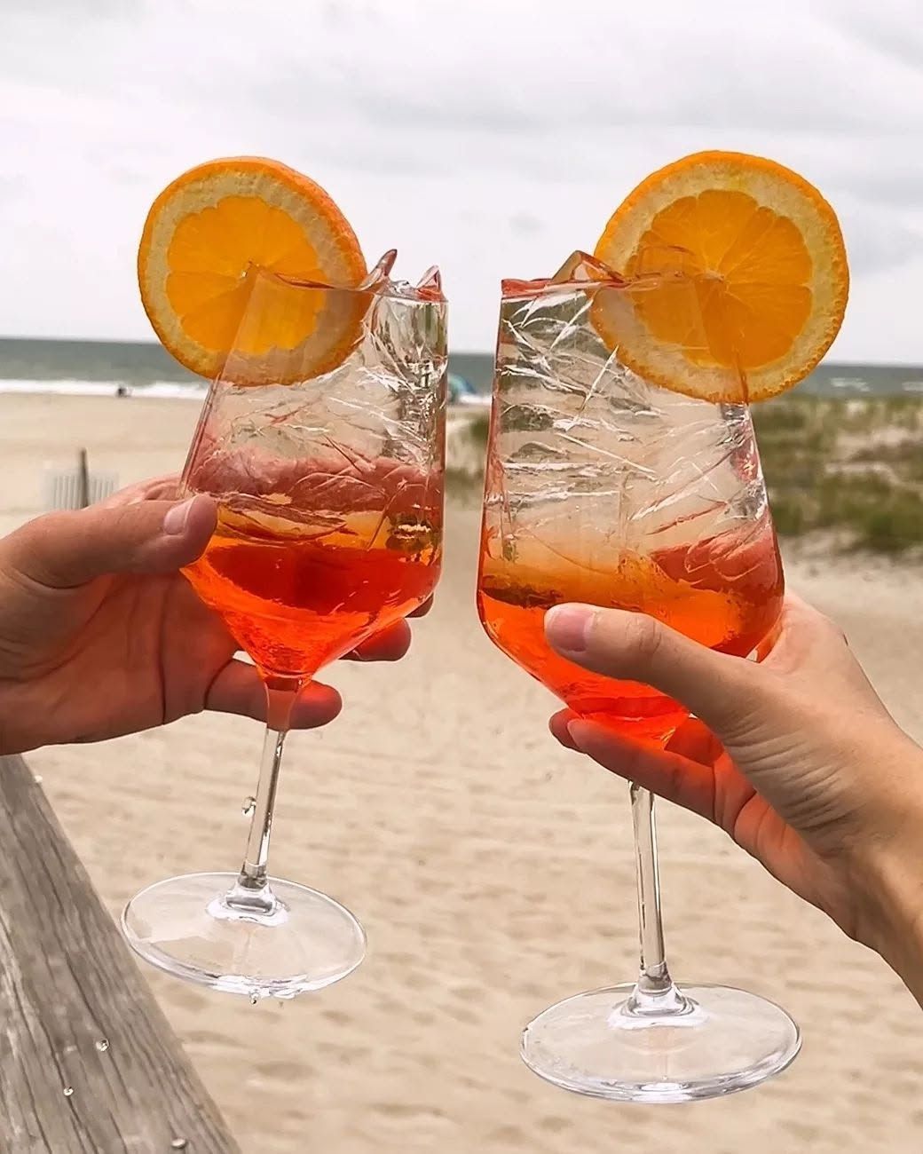 Two hands clinking orange cocktails with orange slices on a beach
