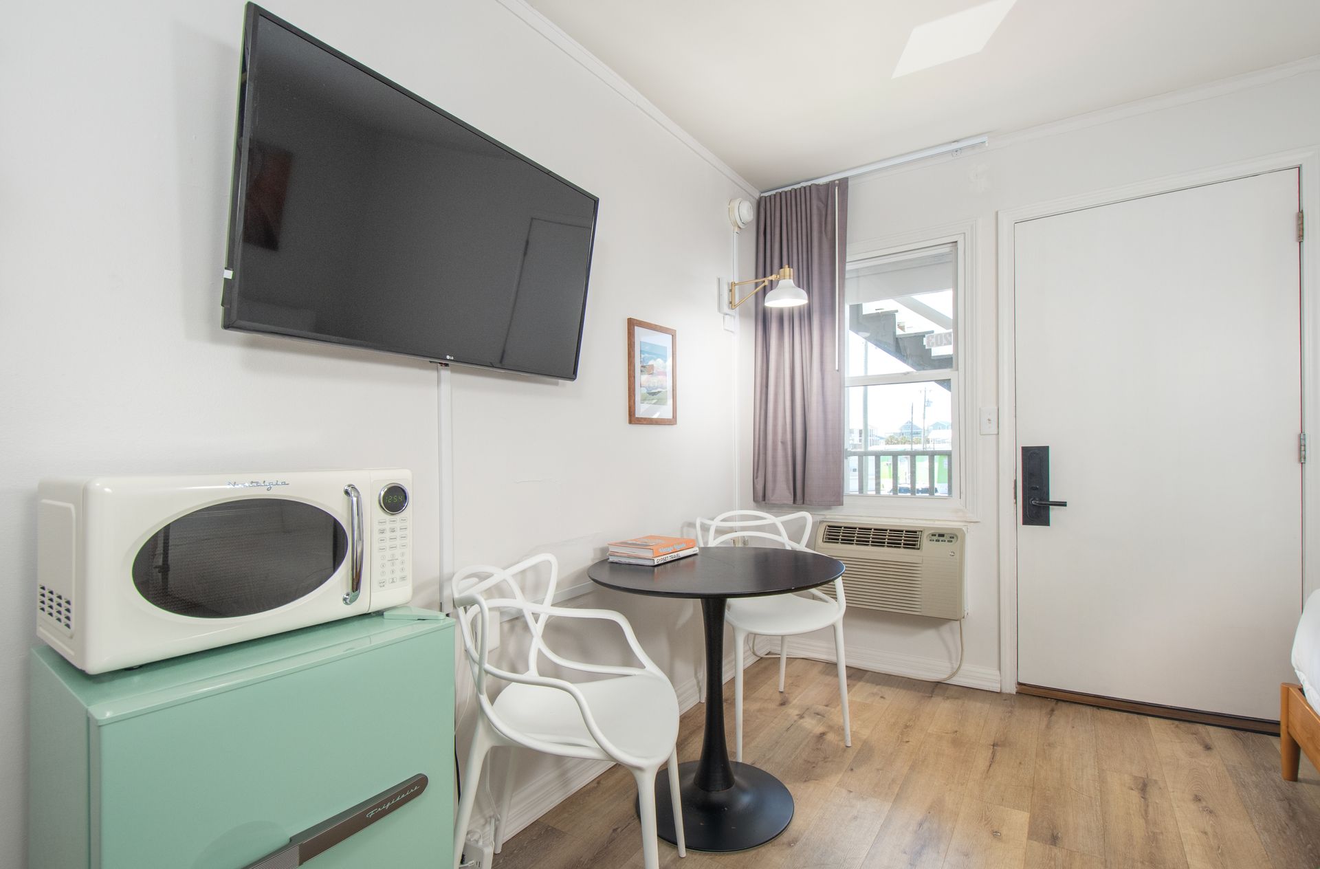 Small room with wall-mounted TV, microwave, table and chairs, mint cabinet, and a door by a window.