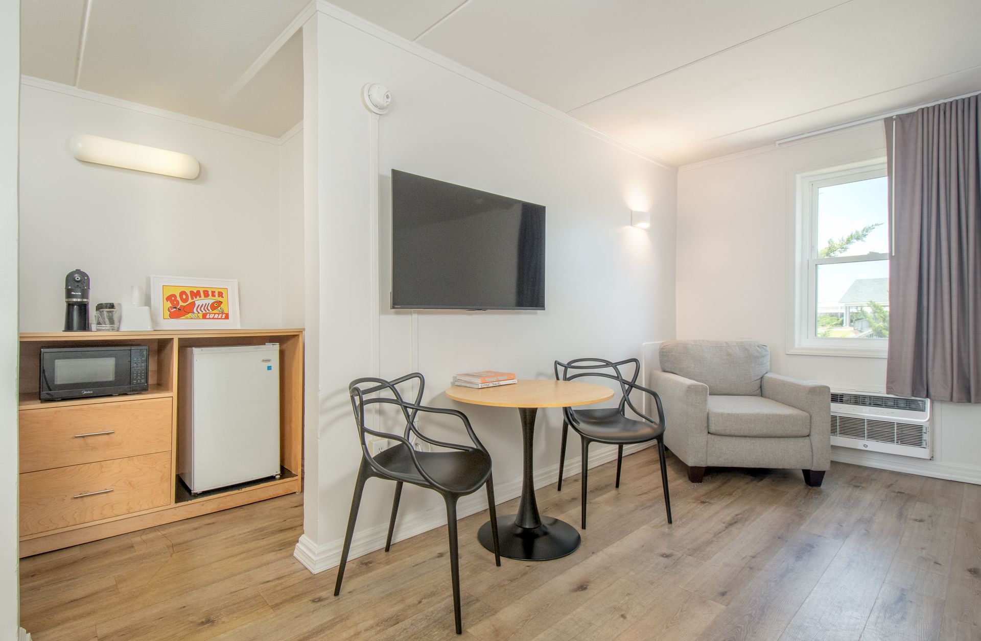 Small studio with a dining table, two chairs, armchair, wall-mounted TV, and kitchenette by a window.
