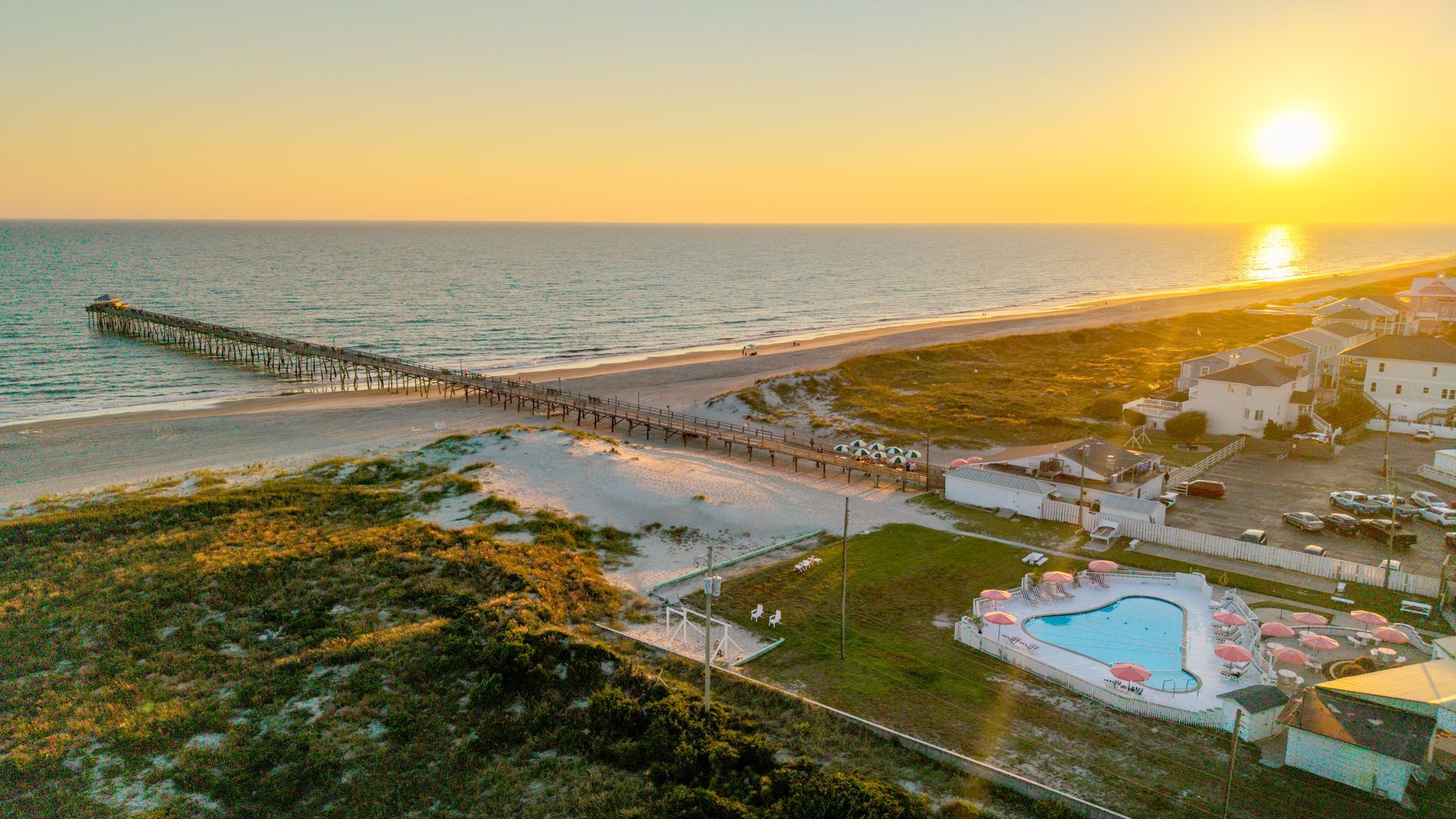 Sunset over a beach resort with a pier, ocean waves, and a pool in the foreground