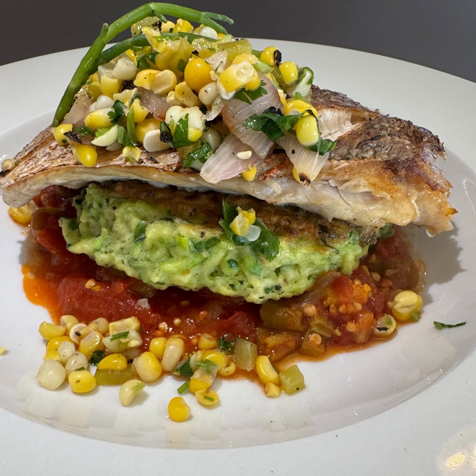 Grilled fish over mashed avocado and tomato sauce, topped with corn salsa on a white plate