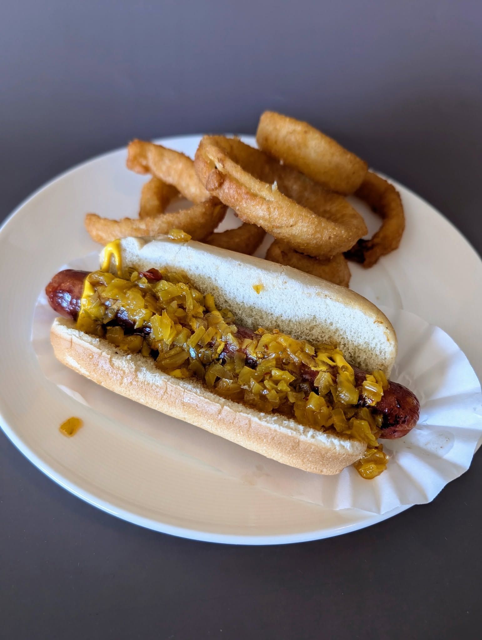 Hot dog with relish on a plate, with thick-cut fries in the background