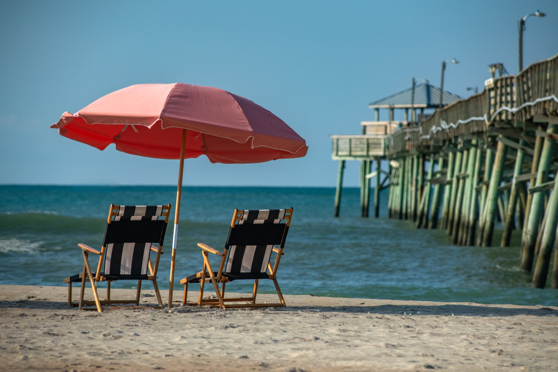 Two beach chairs under a red umbrella beside a pier on a sunny beach