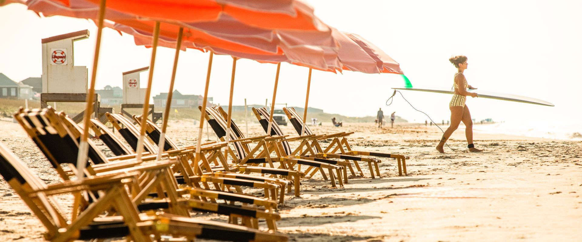 Beach lounge chairs under orange canopies with a person walking along the shoreline carrying a surfboard