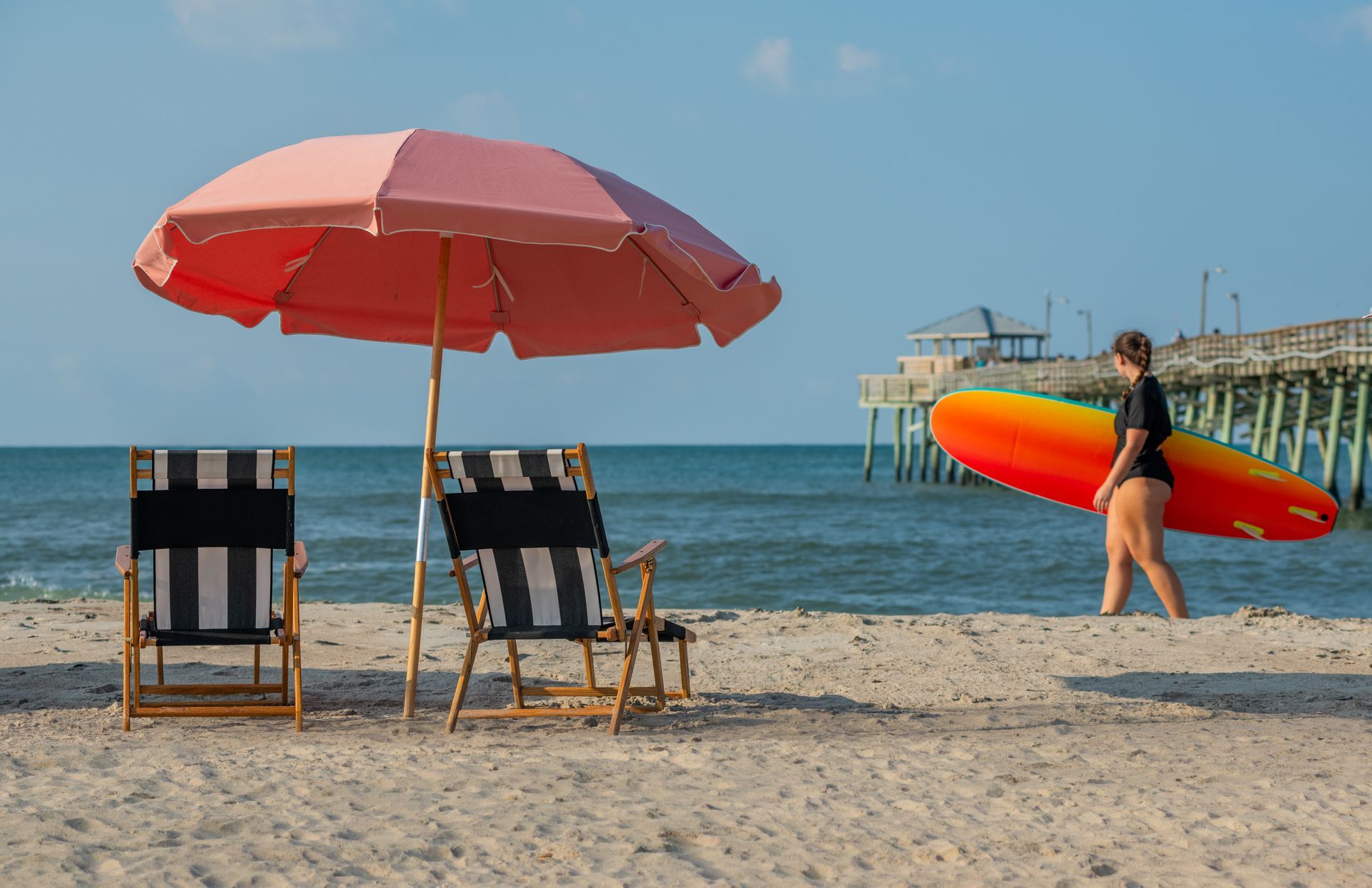 Beach chairs under a pink umbrella with a person carrying a red-orange surfboard by the water