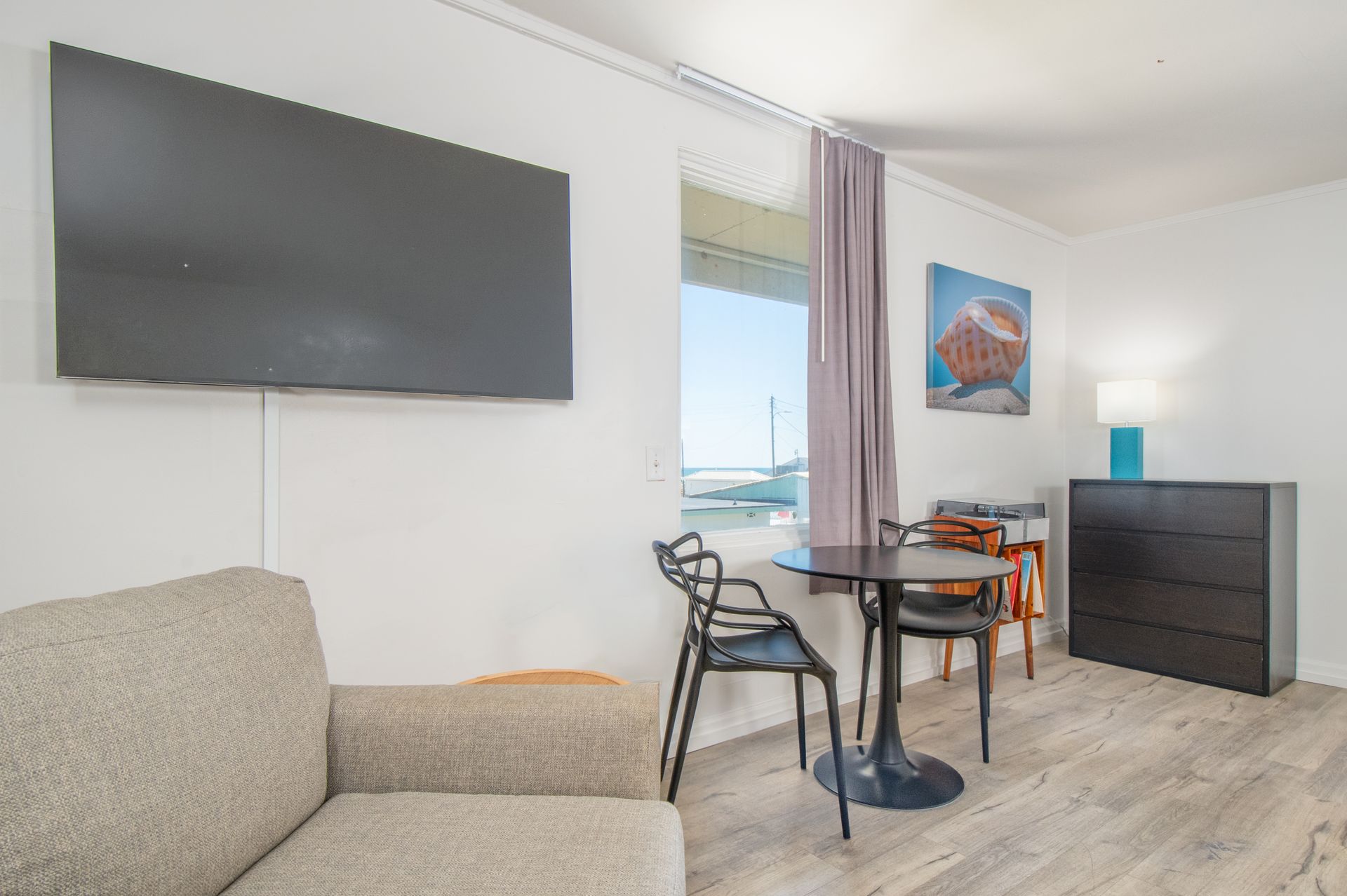 Bright modern room with wall-mounted TV, sofa, small table and chairs, dresser, and ocean-view window.