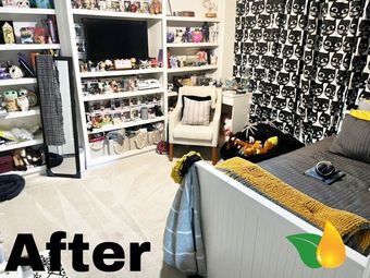 Organized bedroom with shelves of collectibles, a white chair, and a bed beside black patterned curtains.
