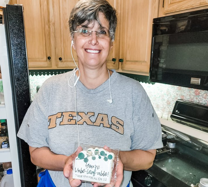 Woman in a kitchen holding a decorated dessert box and smiling, wearing a Texas T-shirt.