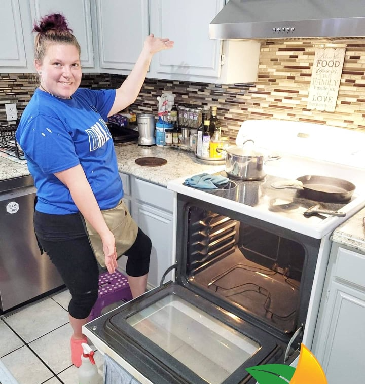 Woman in blue shirt smiling in a kitchen, pointing at an open oven with a casserole inside.