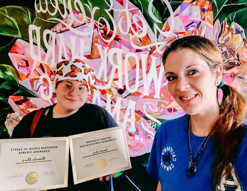 Two smiling people holding certificates in front of a colorful wall mural