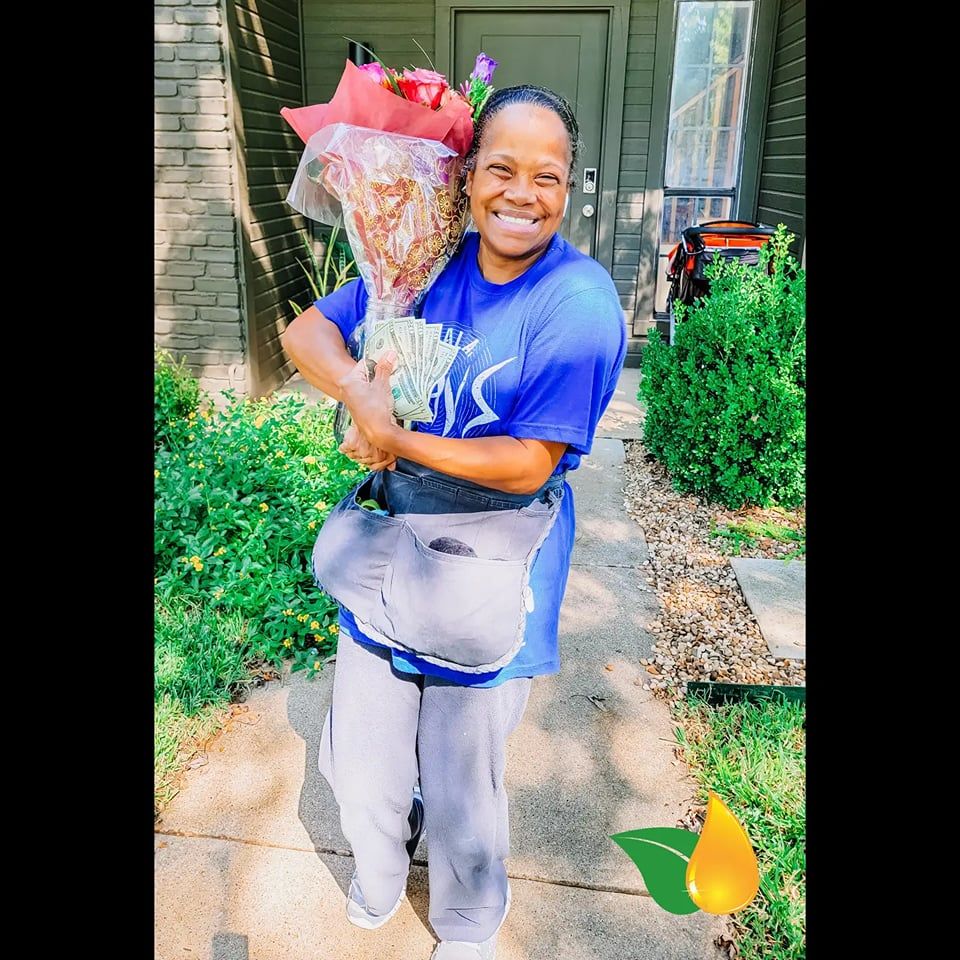Smiling person holding a large wrapped bouquet outside a house, wearing a blue shirt and gray pants.