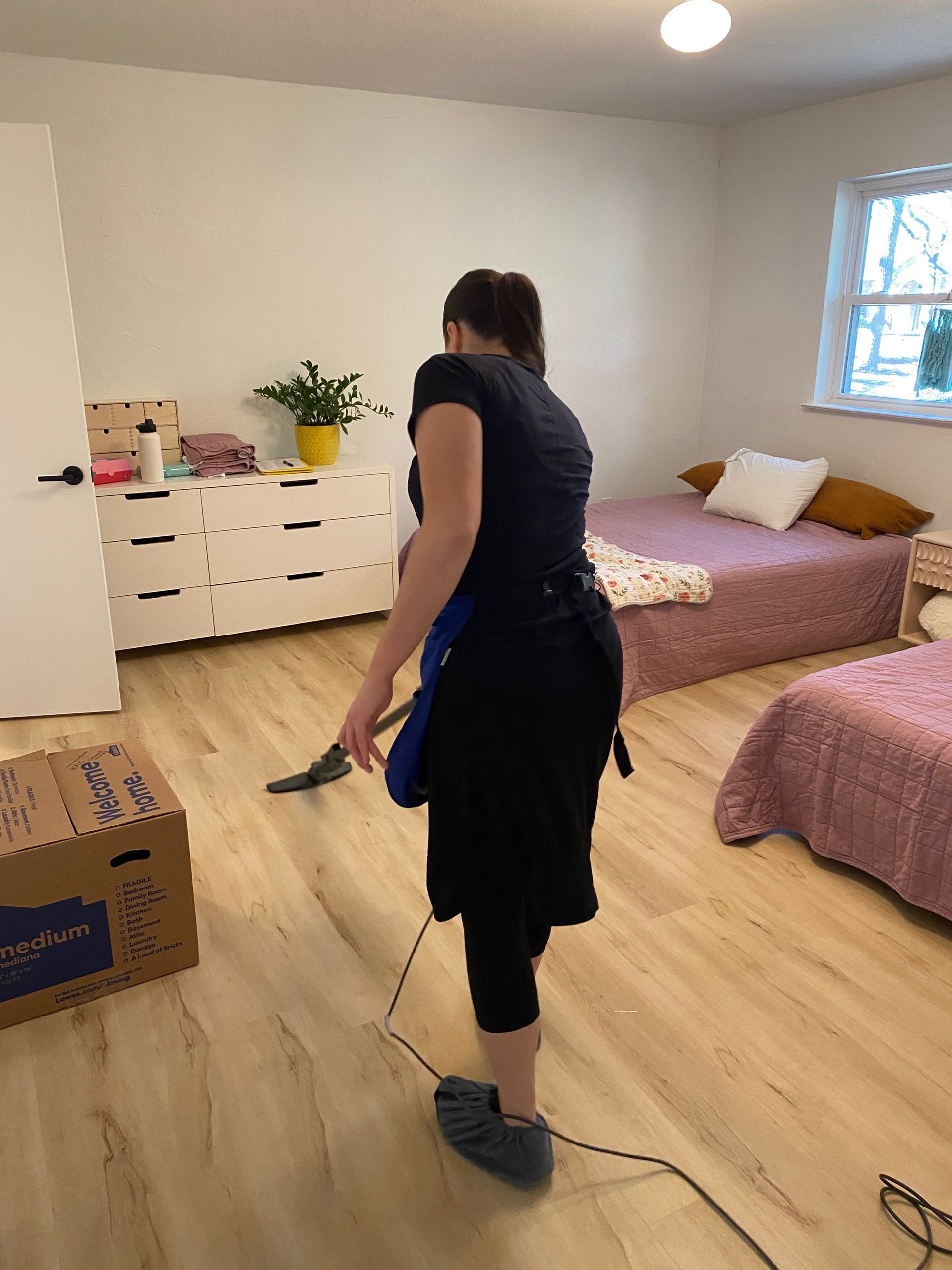 A person cleaning the light wood floor of a bedroom with two beds and a white dresser.