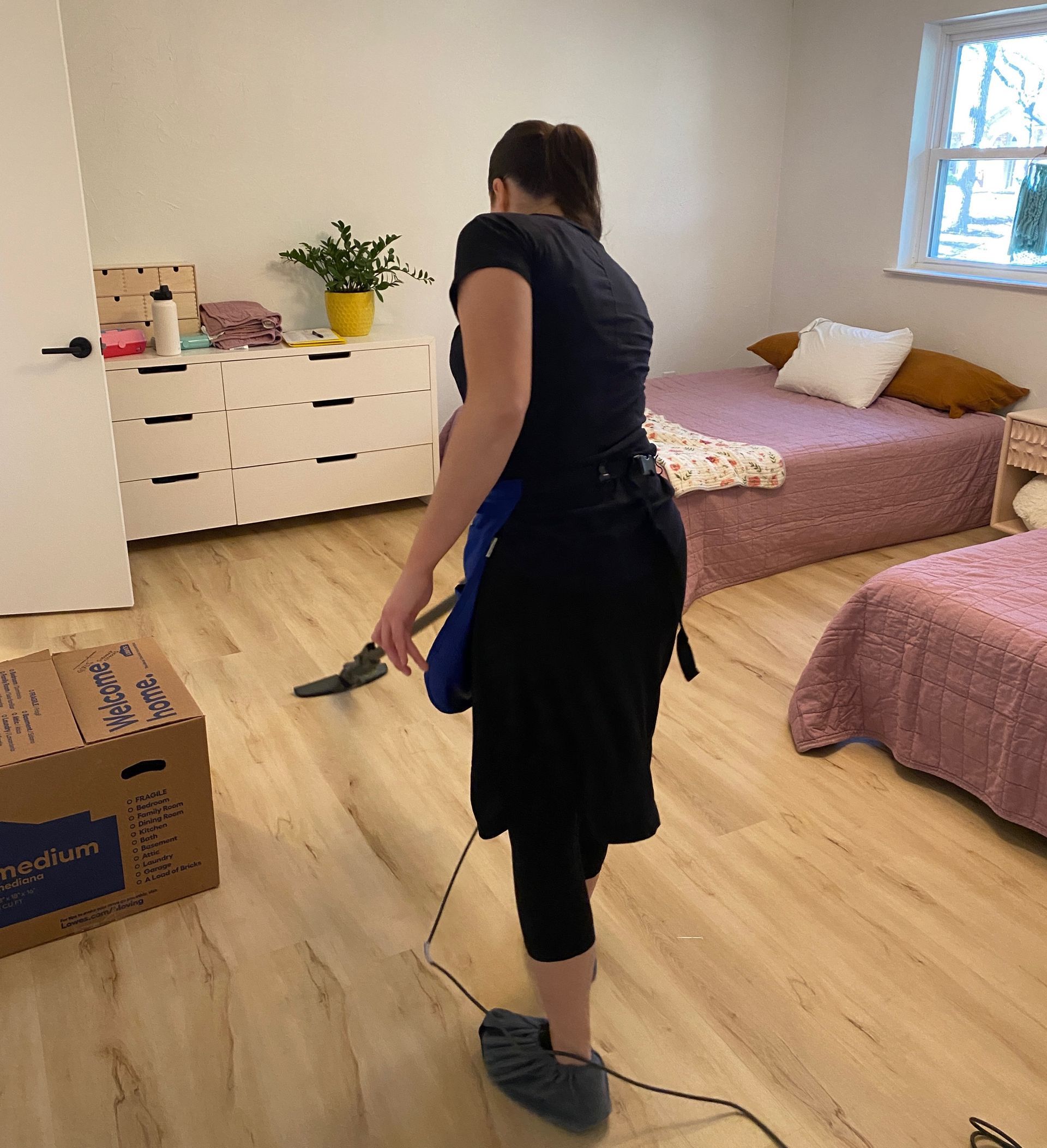 A person vacuums the light wood floor of a bedroom with two beds and a white dresser.