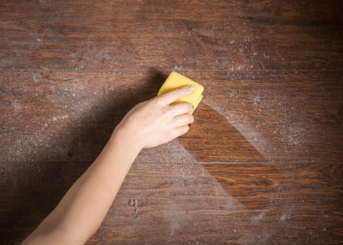 A hand uses a yellow sponge to wipe away a thick layer of dust from a dark wooden surface.
