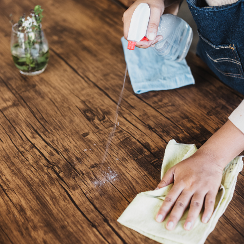 Hands spray a cleaning solution onto a rustic wooden table and wipe it with a yellow cloth.