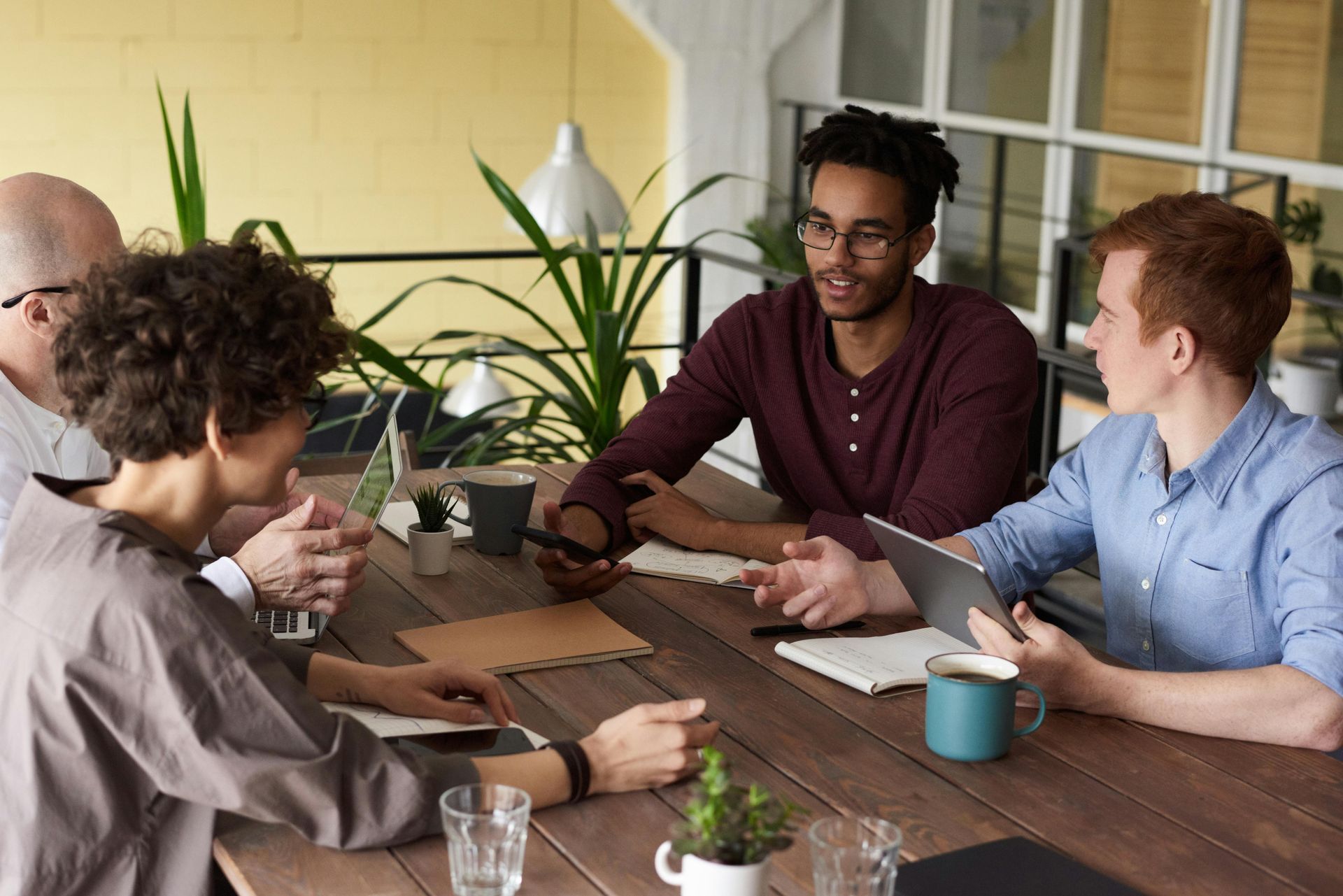 Four people discuss around a wooden table in a bright office with papers, laptops, and coffee mugs.