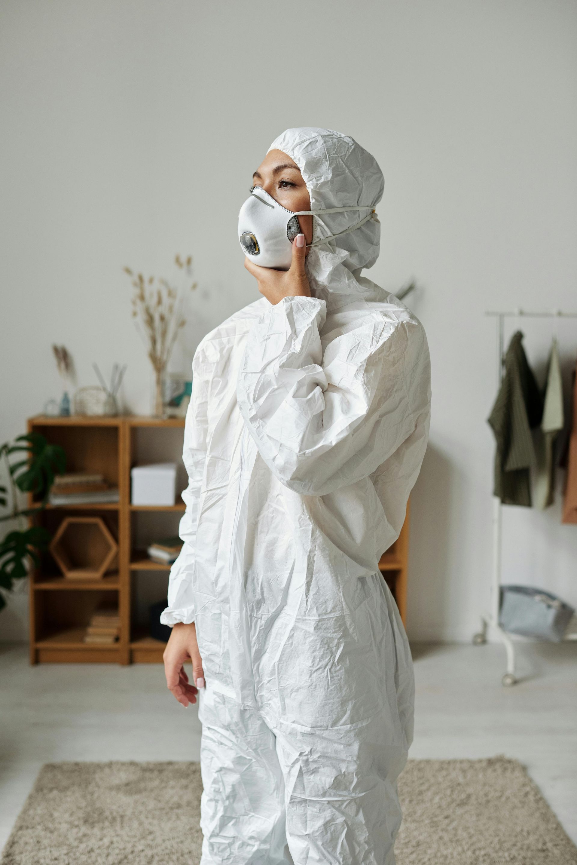 A person in a white hooded protective suit and respirator mask stands in a room with shelving and a clothes rack.