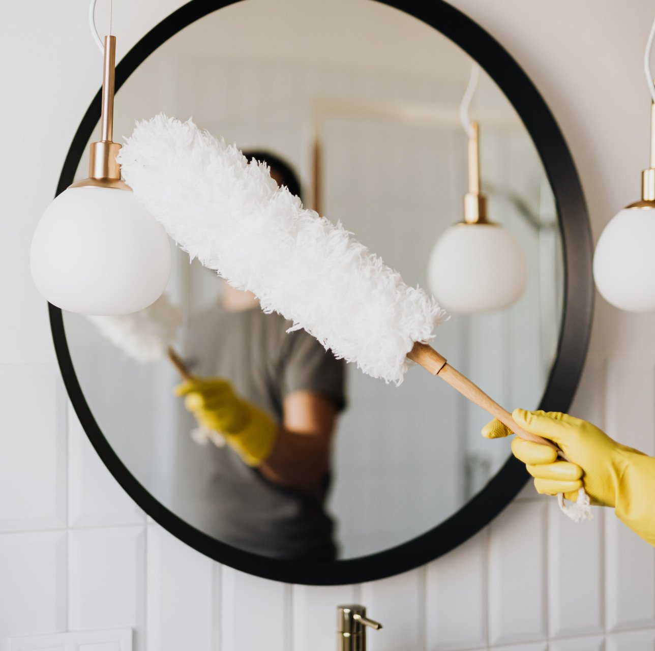 Person dusting a round mirror and wall lamps with a white duster while wearing yellow gloves