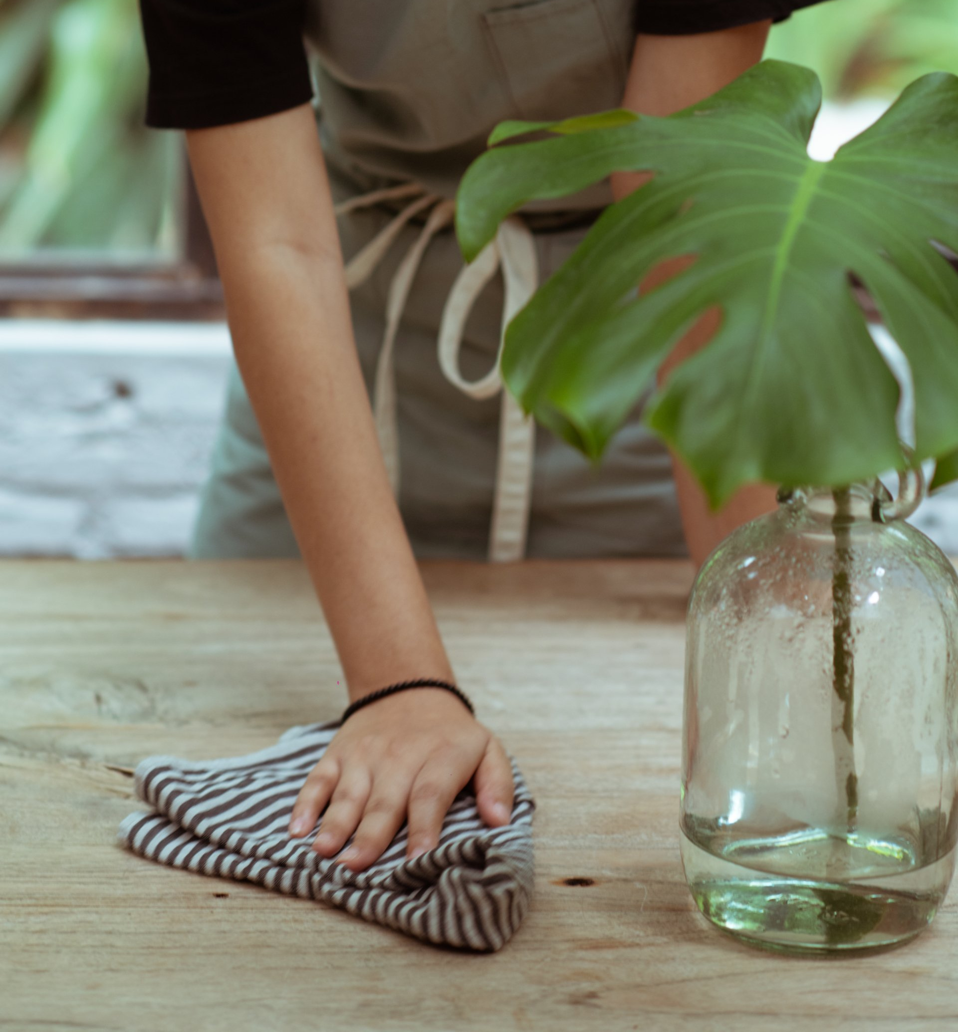 Hand wiping a wooden table with a striped cloth beside a glass vase with green leaves