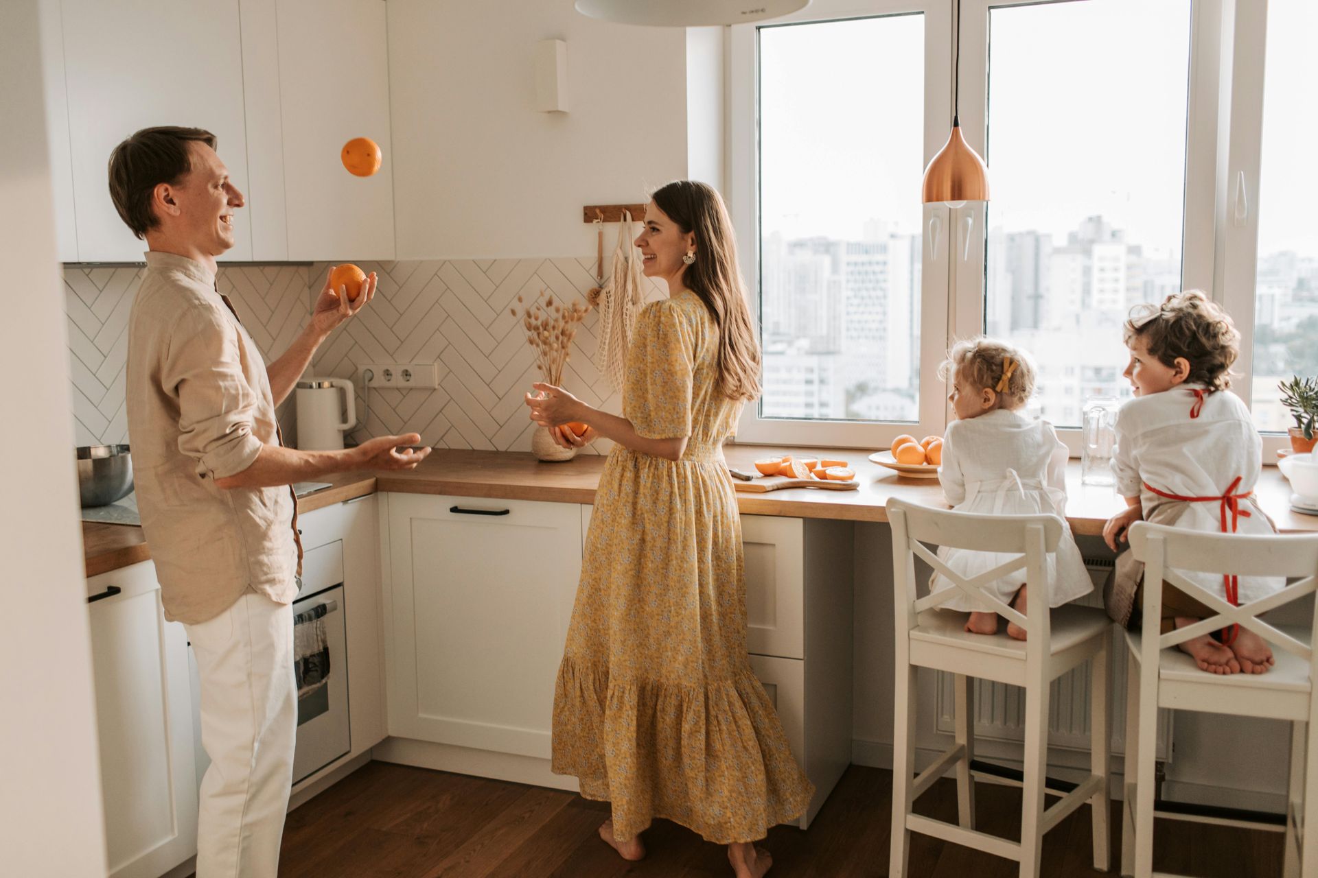 A family in a bright kitchen: a person juggles oranges while others watch and prepare food near a sunlit window.