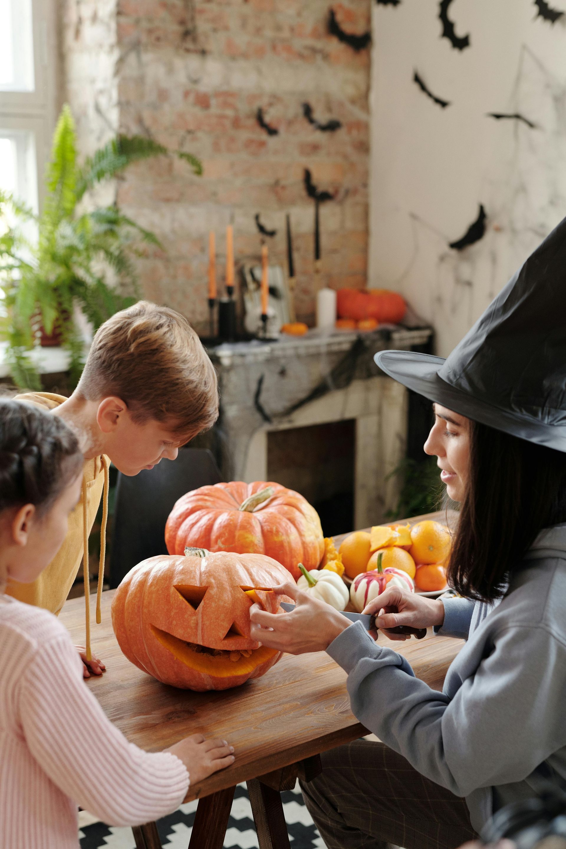 Family carving pumpkins at a table in a cozy kitchen, with Halloween decorations and a witch hat.