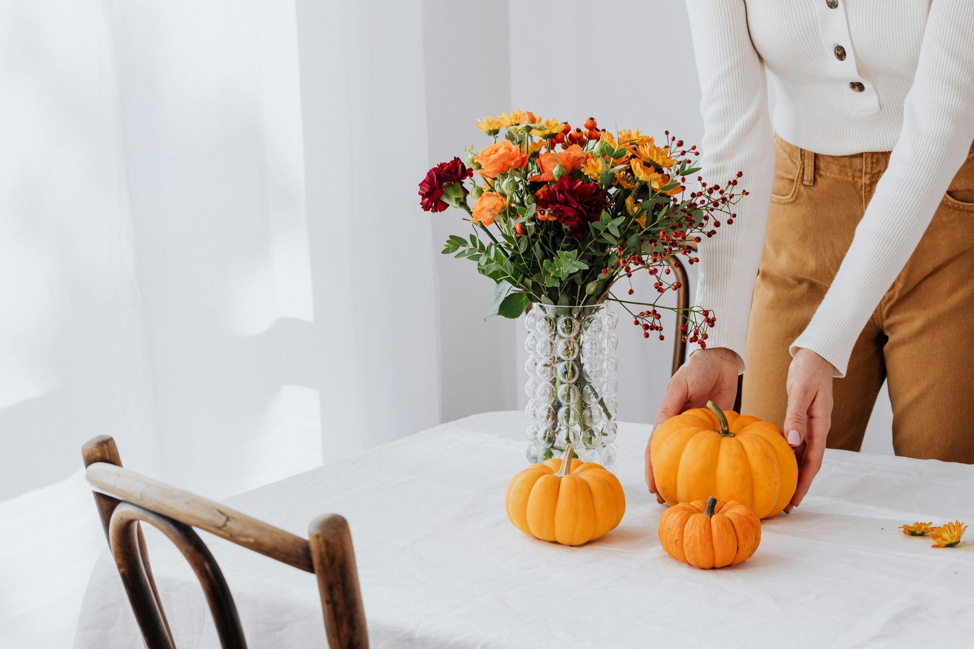A person in a white top and tan pants arranges small pumpkins on a white table next to a vase of autumn-toned flowers.