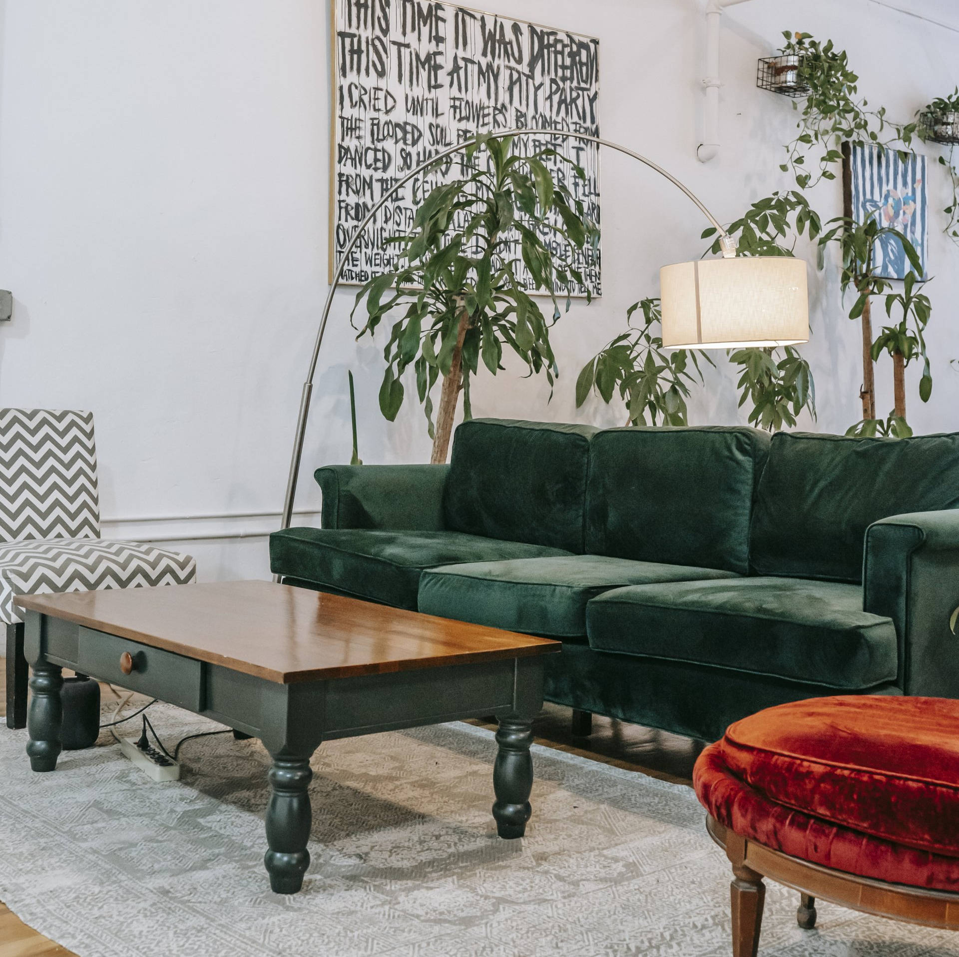 A green velvet sofa and wooden coffee table sit on a patterned rug in a bright, plant-filled living room with art.