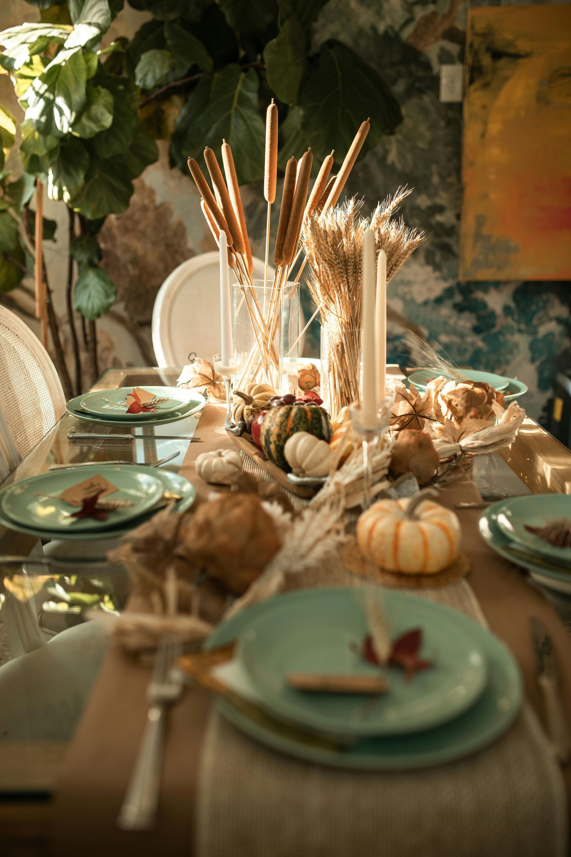 A Thanksgiving-themed dining table with light green plates, small pumpkins, taper candles, and wheat centerpieces.
