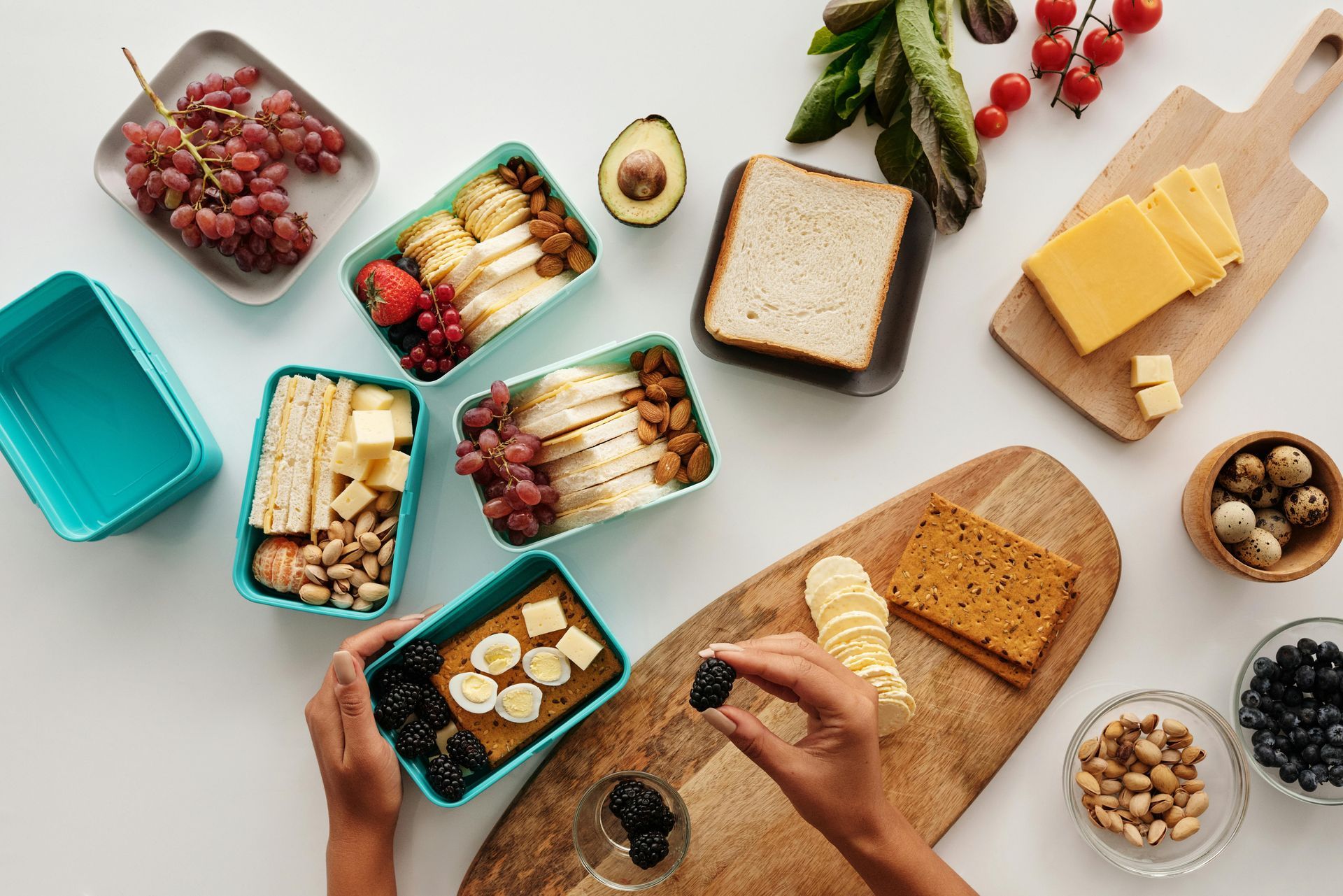 Hands arranging various bento-style meal prep boxes filled with fruit, crackers, and cheese on a white tabletop.