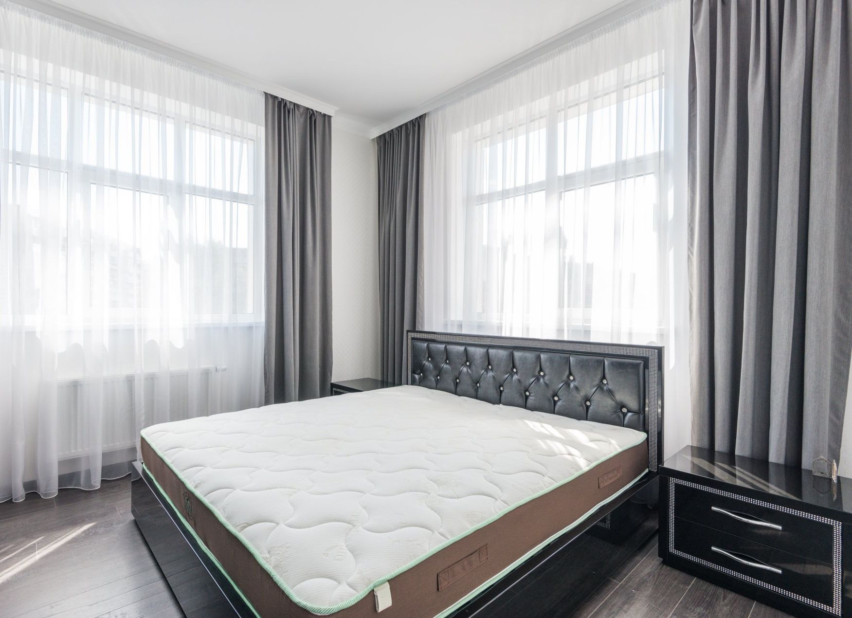A modern bedroom with a tufted black headboard, a white mattress, and gray curtains hanging over bright windows.